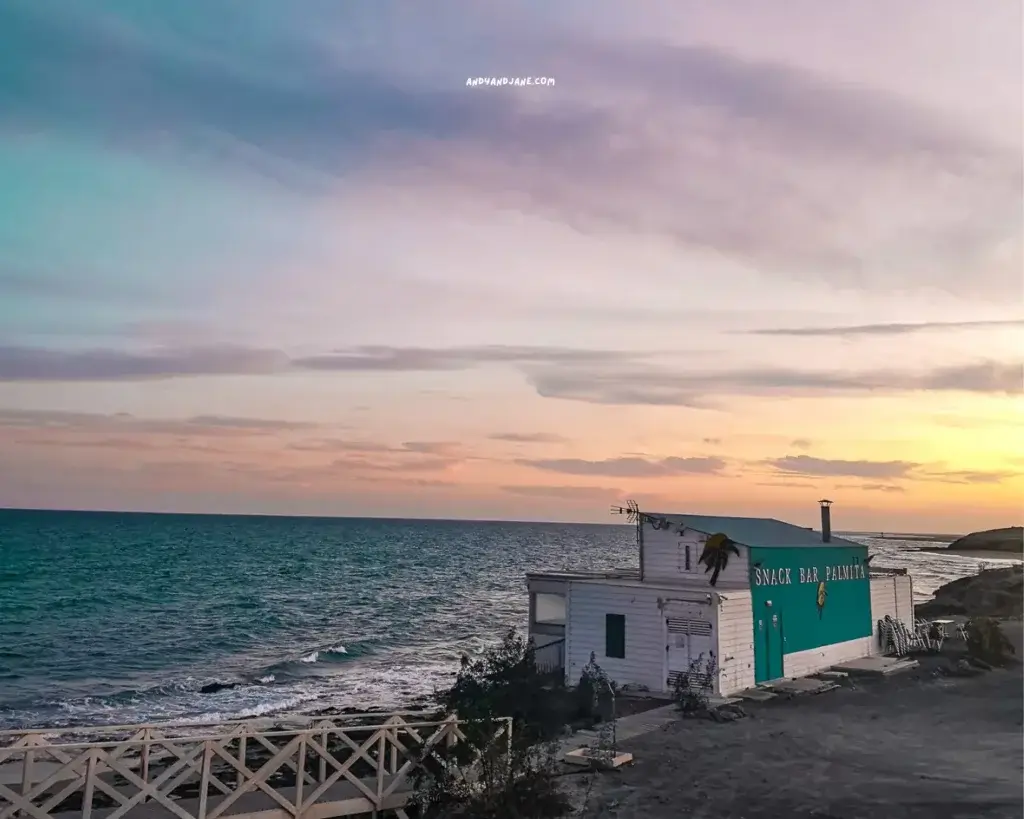 A charming beach bar called Snack Bar Palmita by the sea under a sunset sky with a small white bridge leading to it.