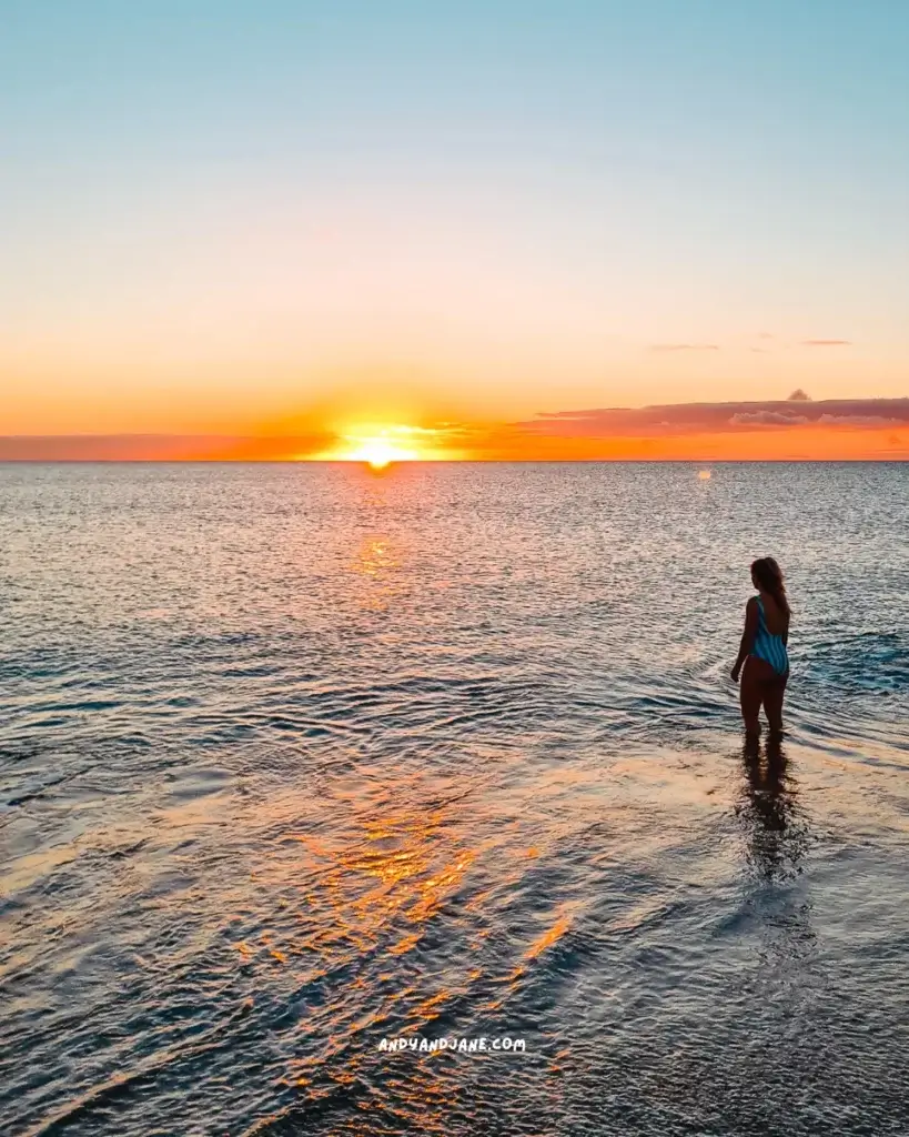 A woman in a green stripe bathing suit standing in the sea watching the sun rise.