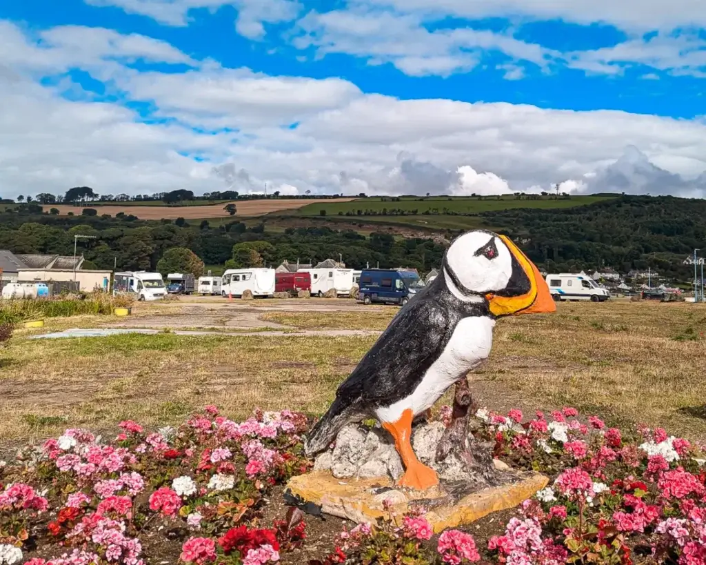 Statue of a puffin surrounded by flowers with a rural landscape and RVs in the background.