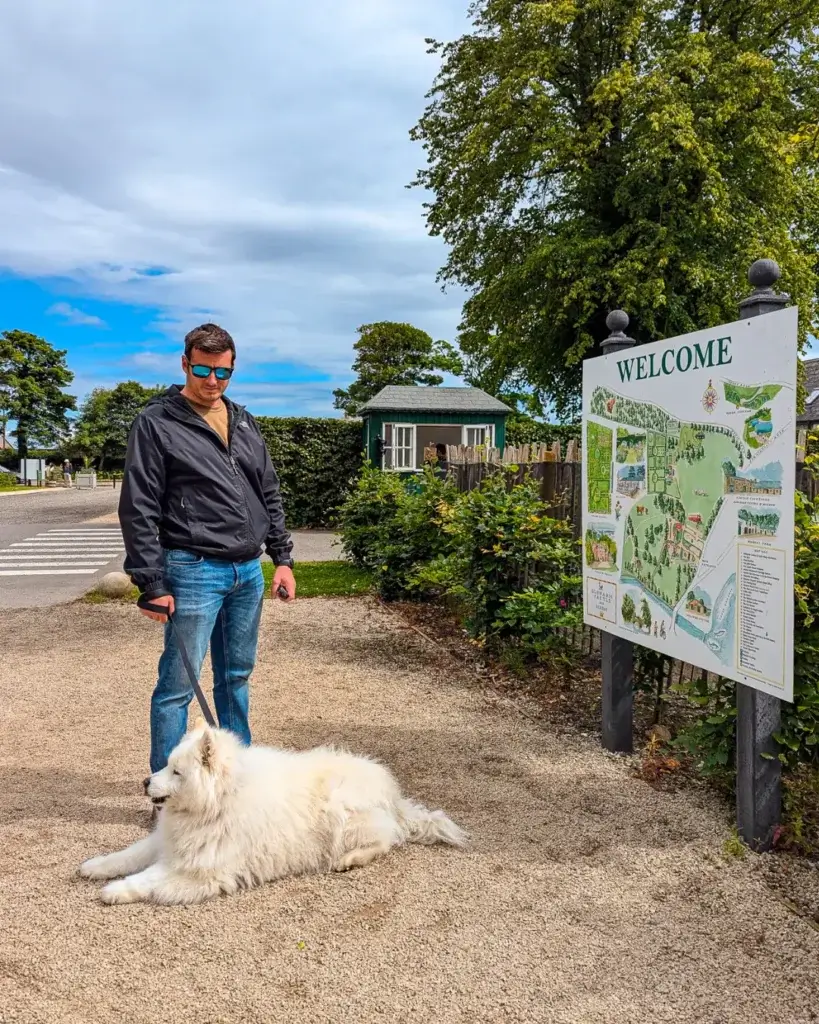 A person with a white fluffy dog by a welcome sign to Glenarm Castle.