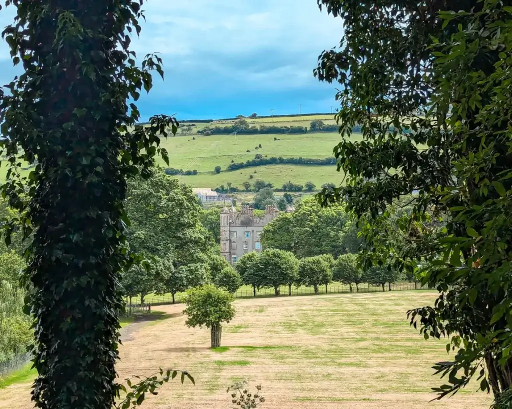 A large, green field framed by trees leads to Glenarm Castle in the distance, with rolling hills in the background under a lightly cloudy sky.