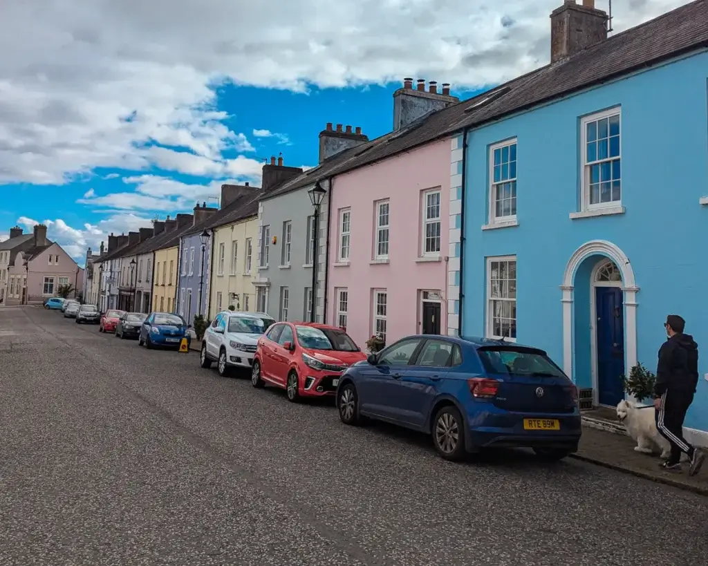 Colorful townhouses line a street in Glenarm with parked cars and a person walking a dog.