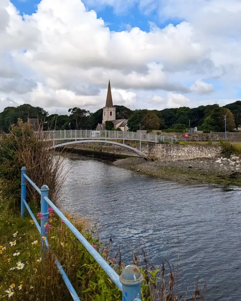 A tranquil scene with a bridge over a river leading to a church spire, under a partly cloudy sky.