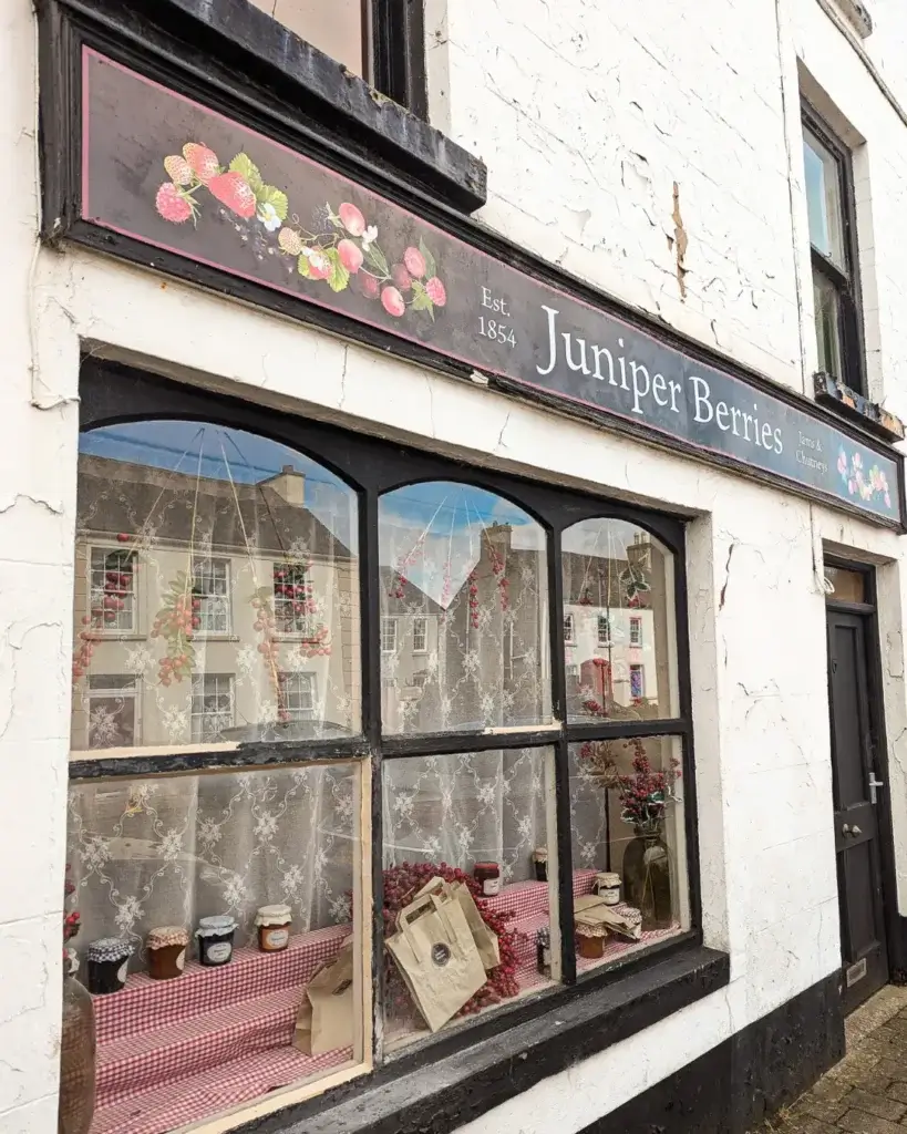 Vintage storefront with a sign "Juniper Berries," lace curtains, and display of jars and berries.