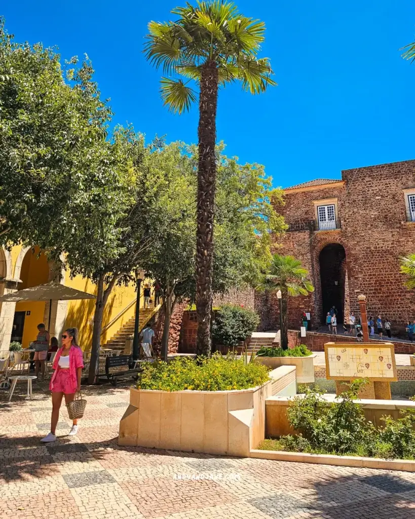 Jane walking through the square in Silves, Portugal - with a palm tree towering over the blue sky & an old city gate in the background.