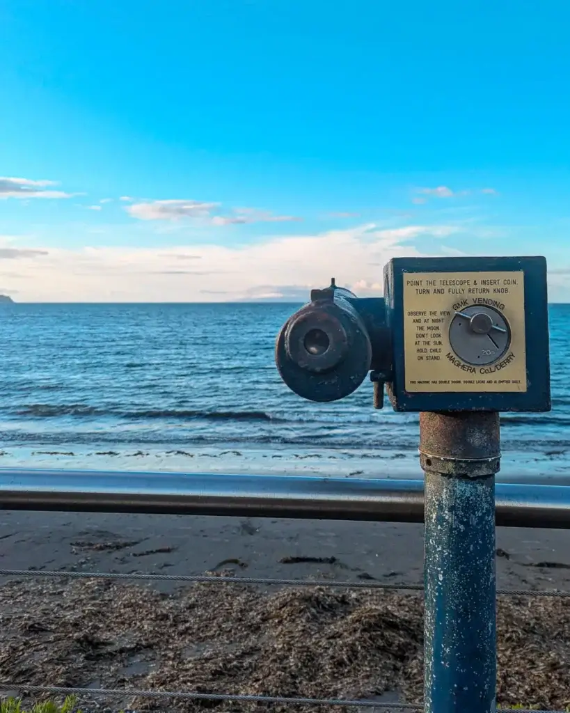 Coin-operated telescope by the seaside with instructions, overlooking calm blue waters under a clear sky.