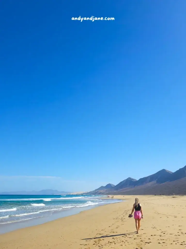 Woman walking on a sunny beach with mountains in the distance and clear blue sky.