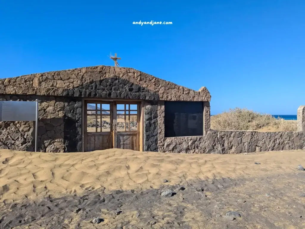 Stone building entrance to a graveyard on the beach with wooden doors on a sandy terrain, under a clear blue sky, near a sea.