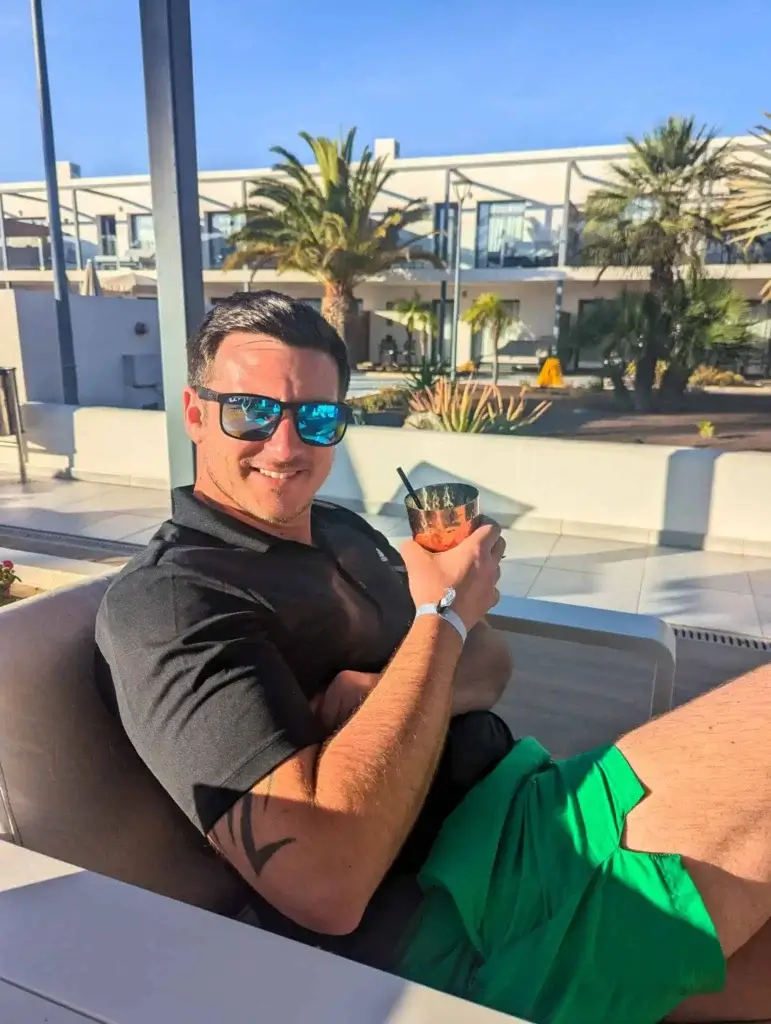 A person relaxes on a lounge chair, holding a drink, wearing a black polo shirt and green shorts, surrounded by palm trees and sunlight.