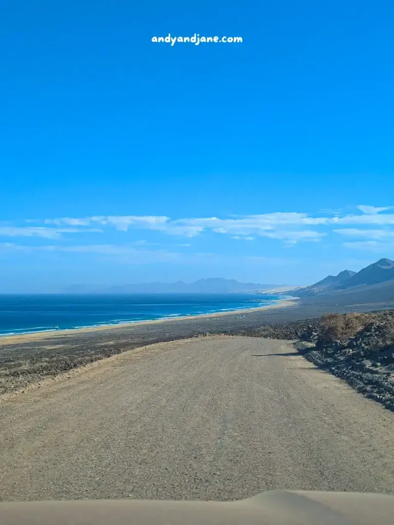 Dirt road leading to a scenic beach with mountains in the distance under a clear blue sky.