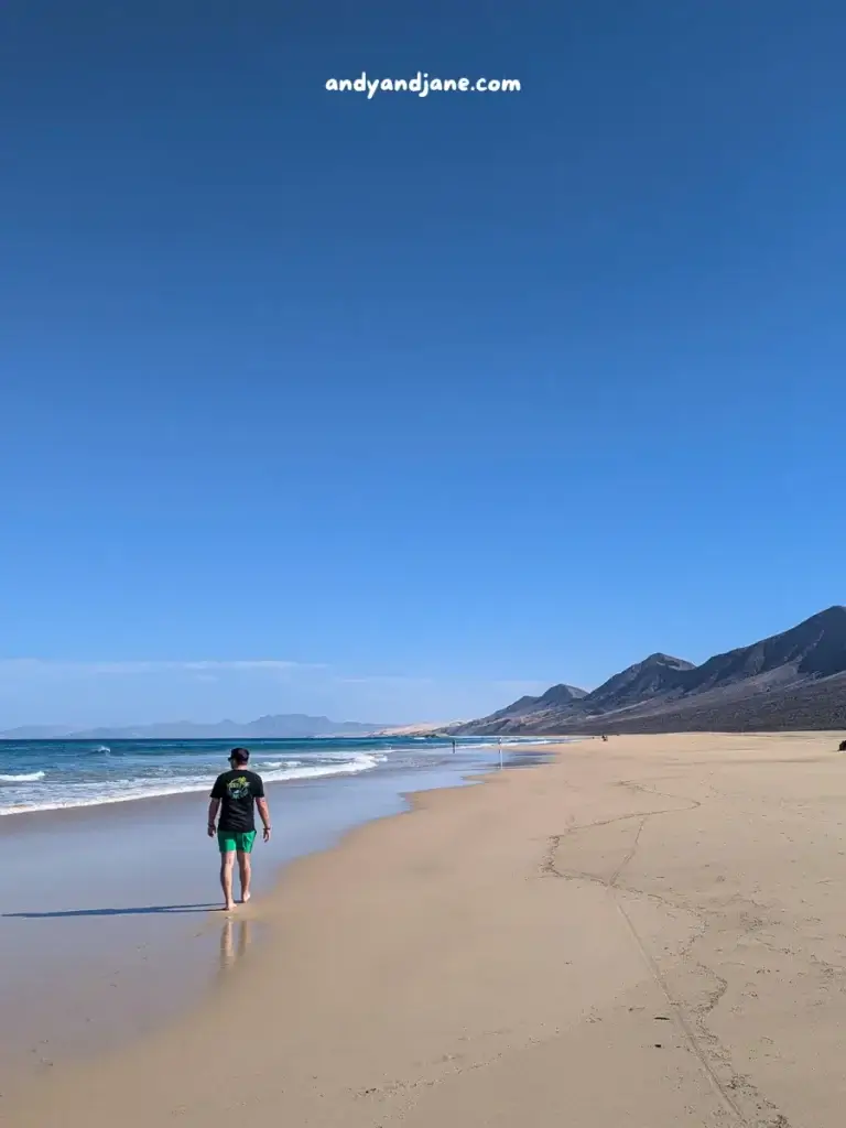 Person walking on the sandy beach of Cofete with clear skies and mountains in the distance.