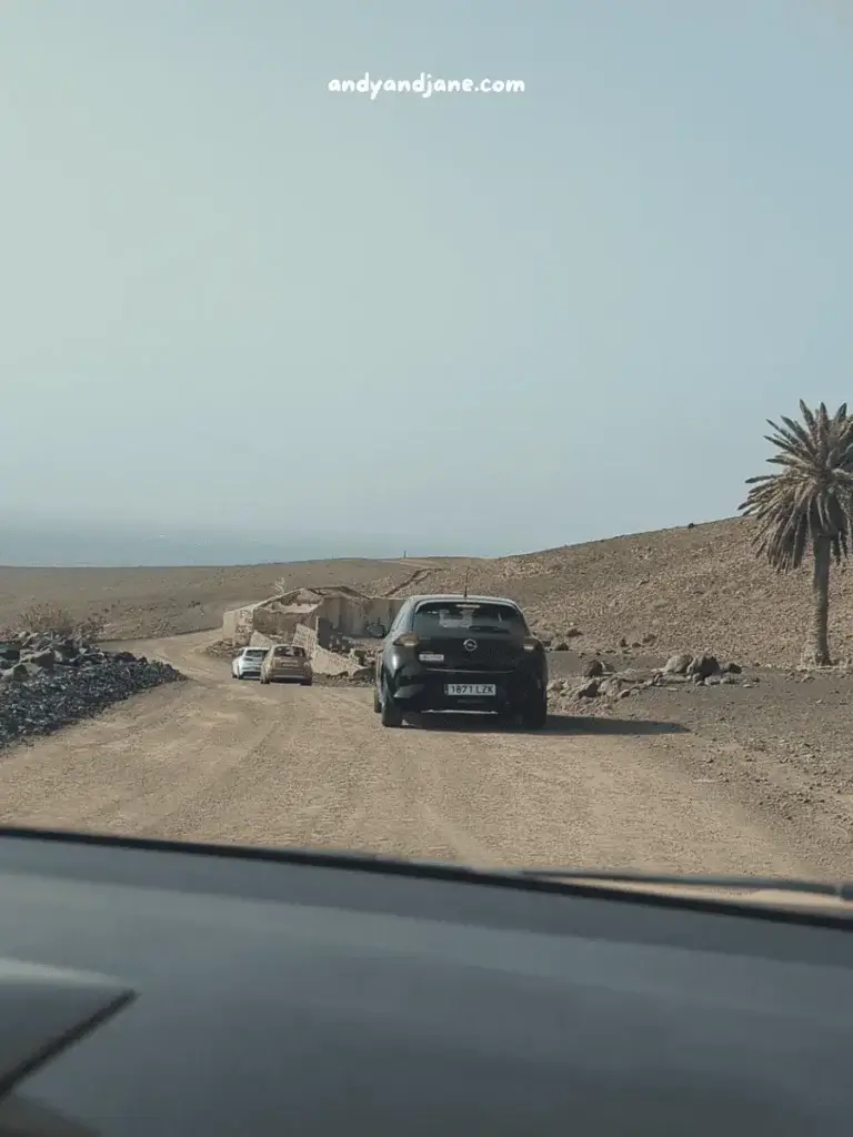 View from a car showing a dusty road with cars and a palm tree, under a clear sky.