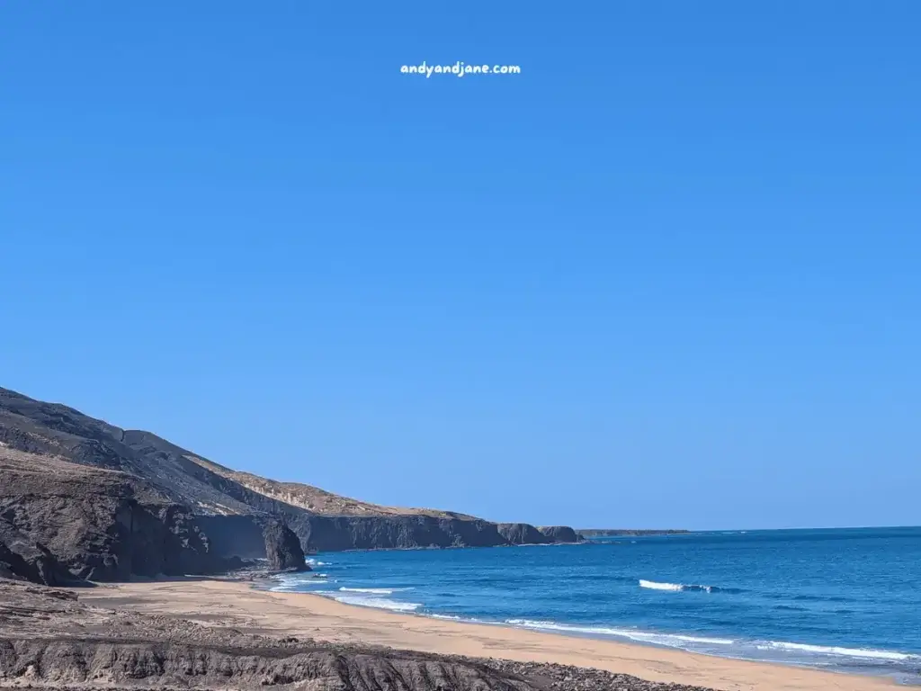 Deserted beach with cliffs under a clear blue sky & a rock formation emerging out of the water at Roque del Morro in Fuerteventura.