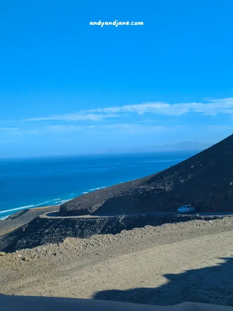 Coastal road winding along a hilly terrain with a blue sea backdrop under a clear sky, and a car parked on the side.