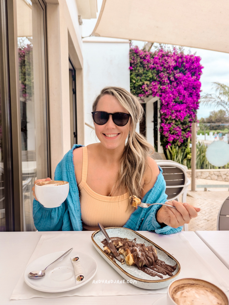 A woman sits at a table outdoors at Doçaria Almeixar, Bistrô, in Vale de Parra Portugal, enjoying a dessert topped with chocolate and bananas, alongside a coffee and a soft drink. 