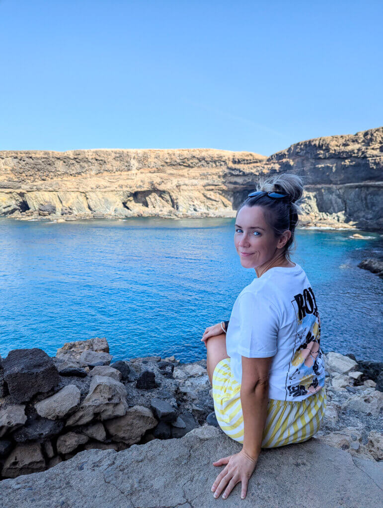 A woman sits on rocky shores, gazing out at calm blue waters and steep cliffs under a clear sky, enjoying a tranquil moment at Ajuy Caves.