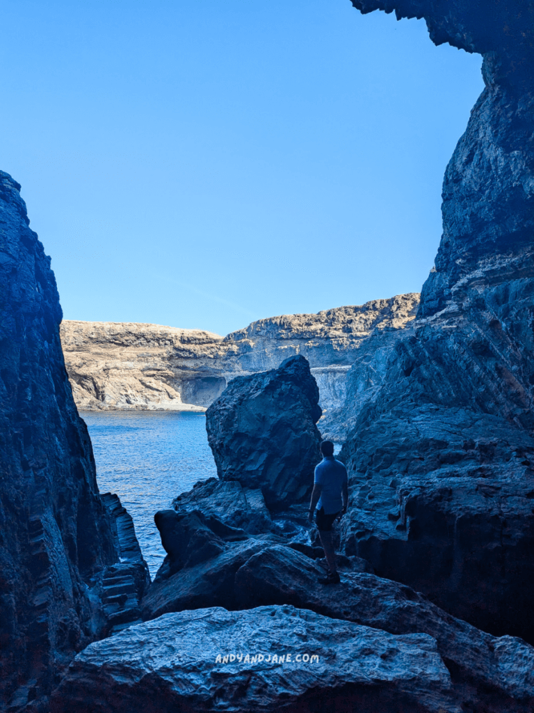 Andrew stands among rugged rocks in the Ajuy caves in Fuerteventura, gazing at the serene blue water and distant cliffs under a clear sky.