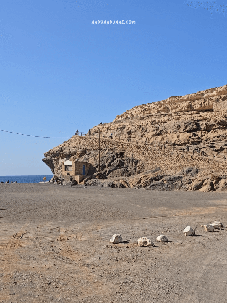 A scenic beach at Ajuy with a rocky cliffside leading up to the Ajuy Caves, a small building at the shore, and visitors exploring the area under a clear blue sky.