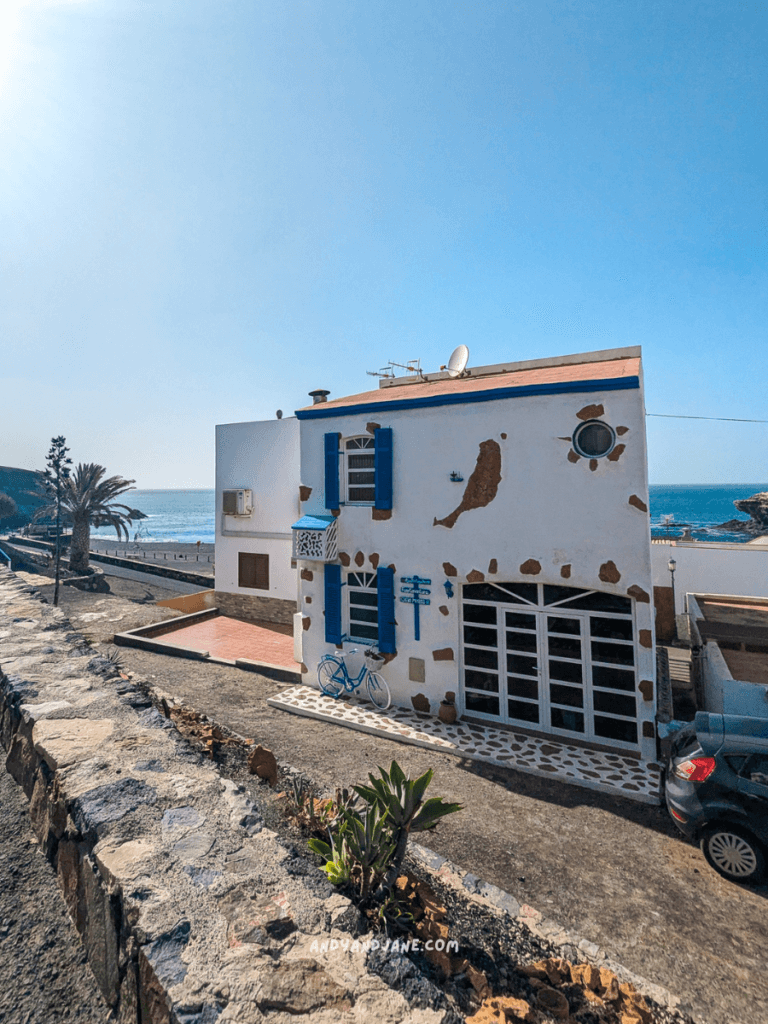 Coastal view of a charming white house with blue shutters, near a rocky pathway and palm trees, overlooking the sea in Ajuy.