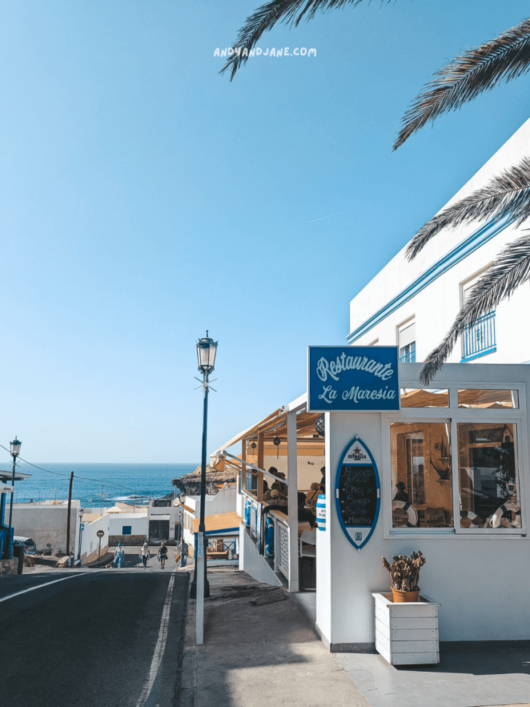 A street scene in Ajuy, Fuerteventura featuring the seaside restaurant "La Maresía," framed by palm trees and ocean views under a clear blue sky.