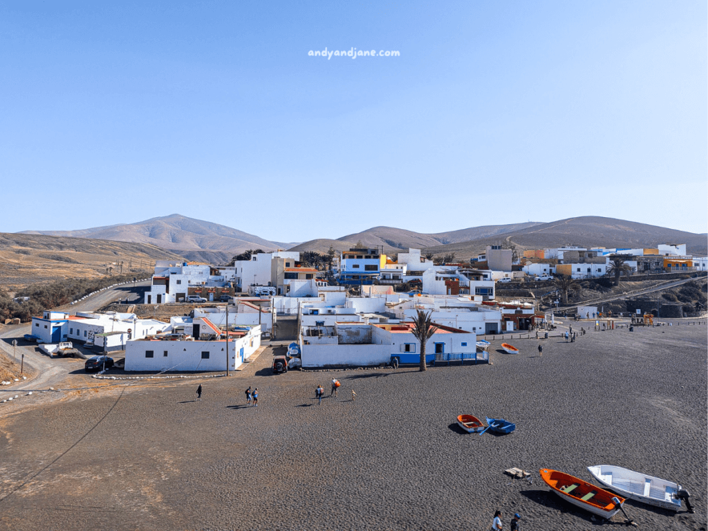 A scenic view of the coastal village Ajuy in Fuerteventura, with white buildings, colorful boats, and surrounding hills under a clear blue sky.