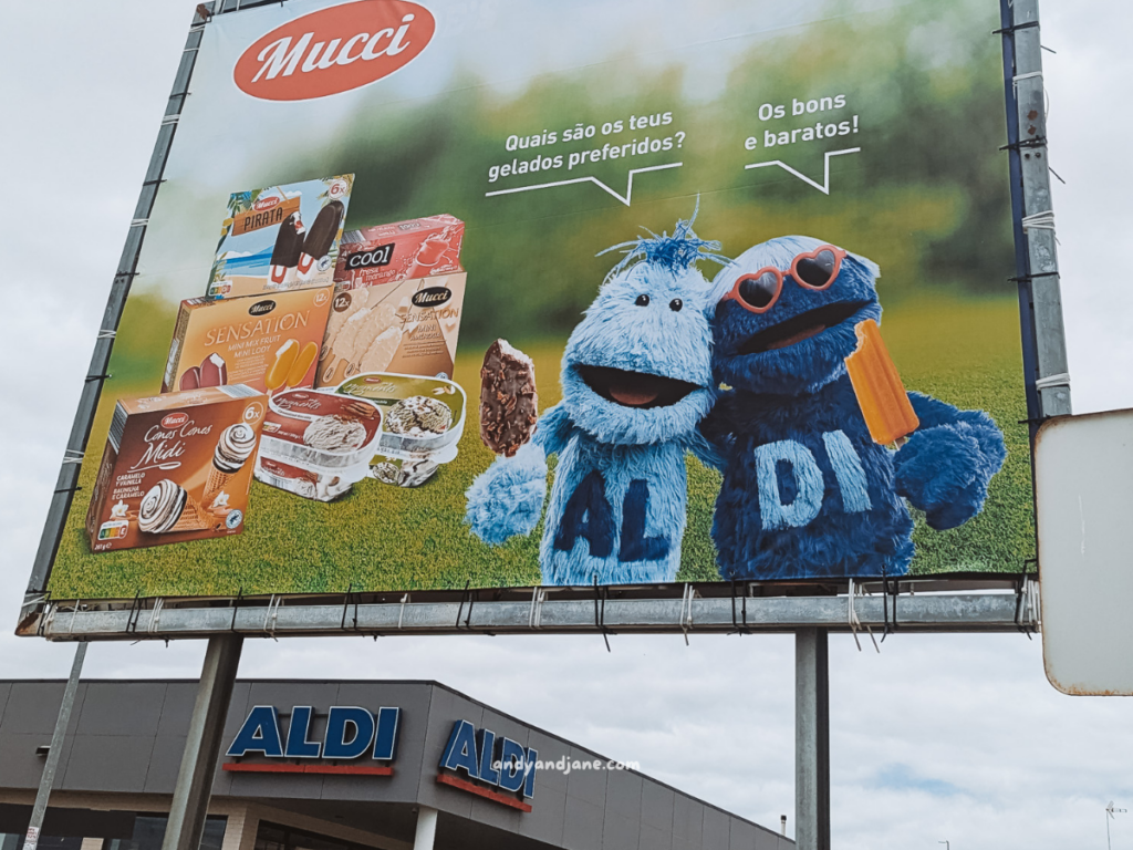 A colorful billboard features two furry blue mascots holding ice cream, promoting Mucci ice creams at Aldi with the slogan, "Good and affordable!"