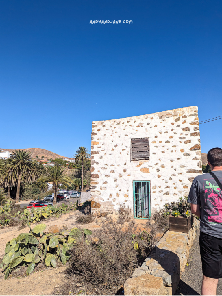 A rocky, white-walled structure with a barred window, surrounded by palms and cacti, against a clear blue sky. A person walks nearby.