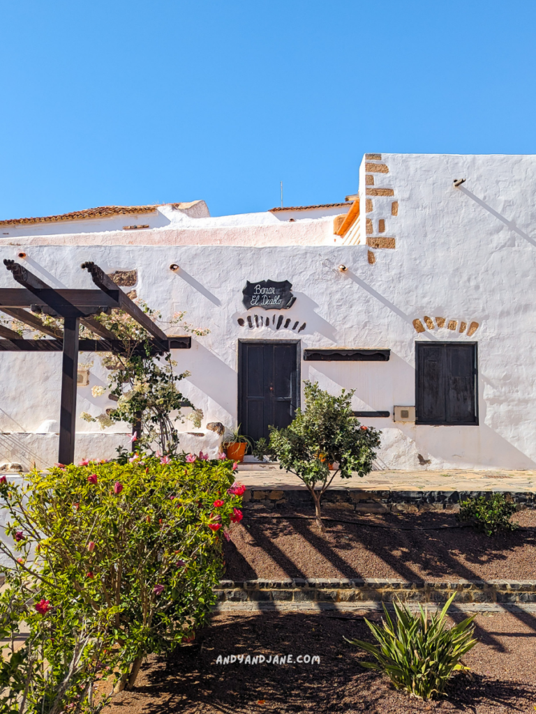 Nestled in Betancuria, this white stucco building features classic wooden doors and is beautifully surrounded by lush plants beneath a clear blue sky.