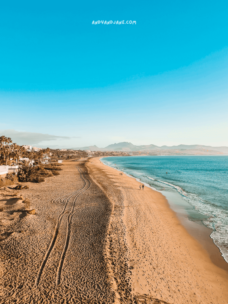 Costa Calma beach with golden sand, turquoise waves, and distant mountains, featuring people walking along the shoreline under a clear blue sky.