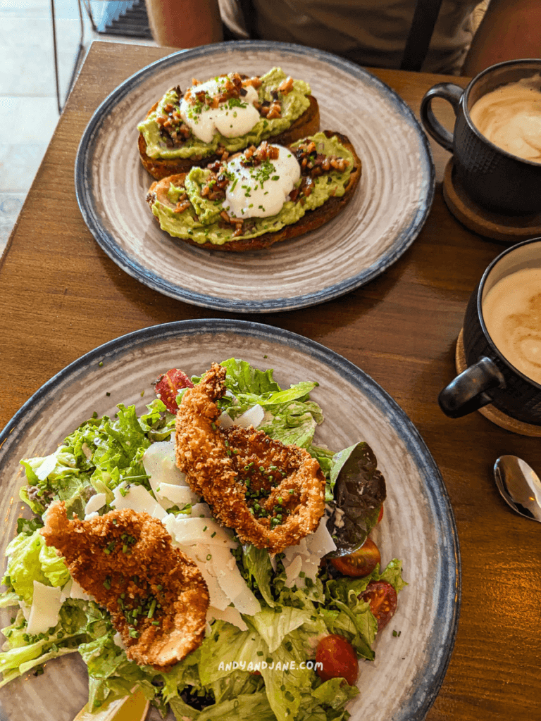 A brunch spread featuring avocado toast with poached eggs and crispy bacon, alongside a salad with fried elements and cheese in Almirante Brunch, Lajares.