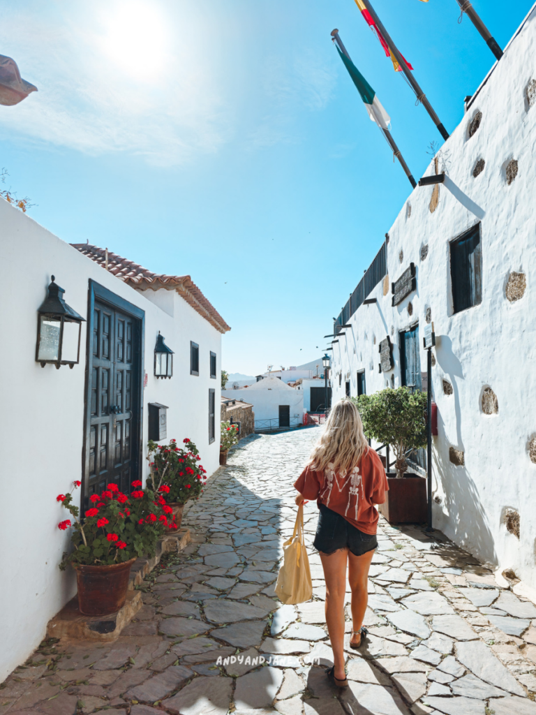 A woman with long hair walks down a sunlit cobblestone street in Betancuria lined with white buildings and vibrant flowers.