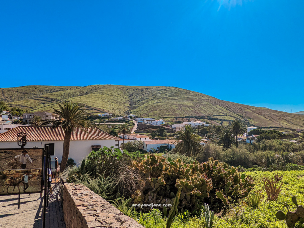 Scenic view of a bright blue sky over green hills and lush vegetation, with a stone wall and houses in the distance.