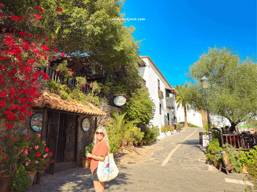 A sunlit cobblestone pathway in Betancuria, Fuerteventura lined with colorful flowers, rustic buildings, and lush trees under a clear blue sky.