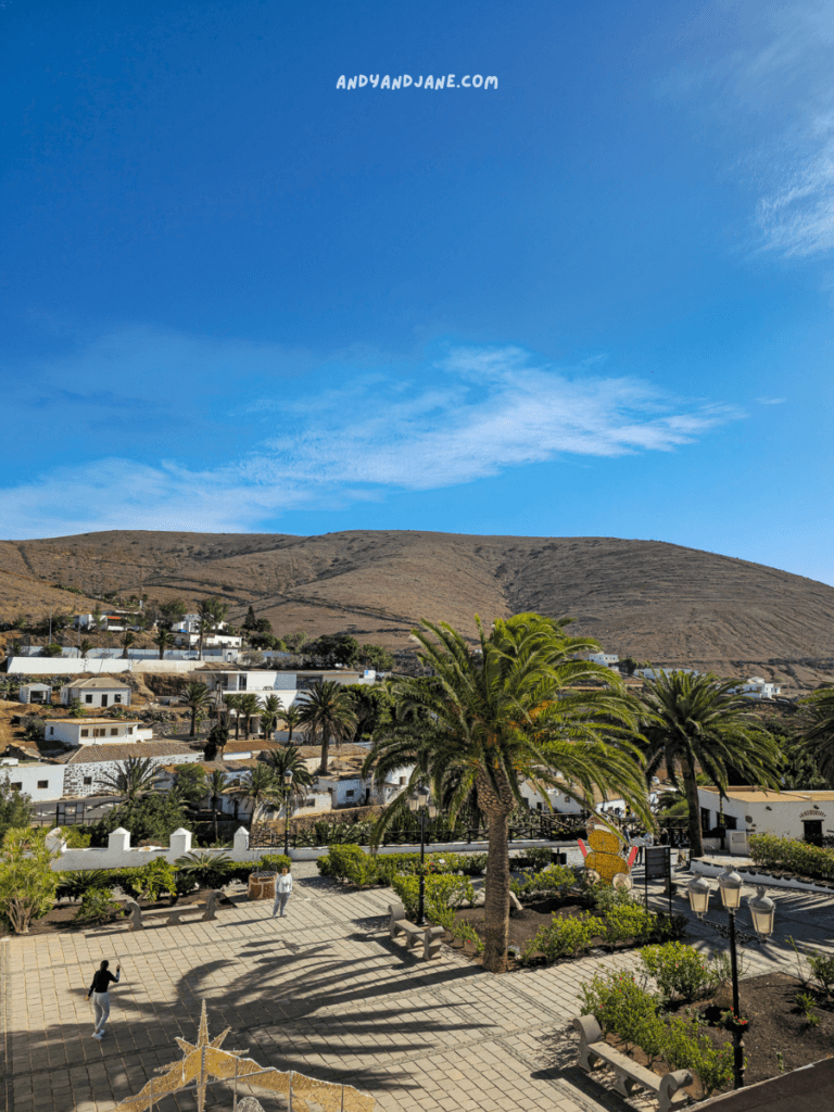 A scenic view of a tranquil village called Betancuria, with palm trees and mountains under a clear blue sky, showcasing a peaceful outdoor plaza.