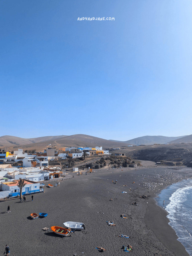 A coastal view of the black sandy beach in Ajuy, Fuerteventura, featuring colorful boats, and people lounging, with hills and colorful buildings in the background.