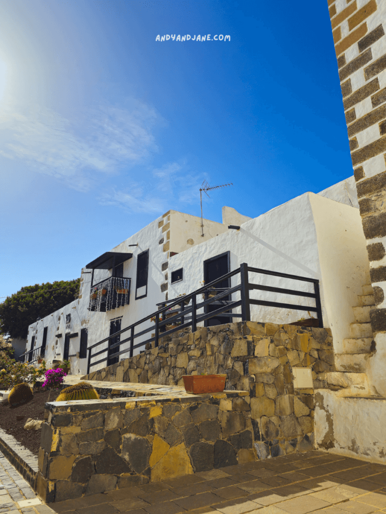 Bright white buildings with black accents, stone pathways, and vibrant flowers under a clear blue sky create a charming residential scene.