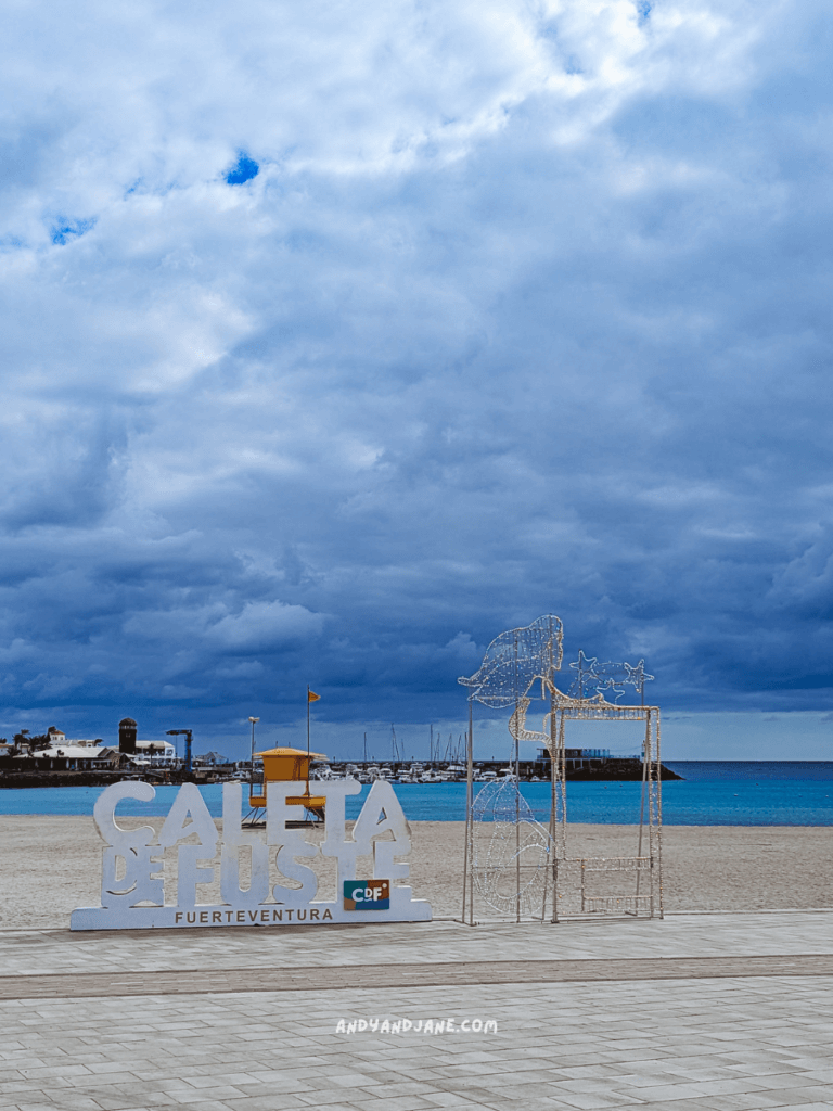 Sign for Caleta de Fuste, Fuerteventura, with a beach and harbor in the background under a cloudy sky.