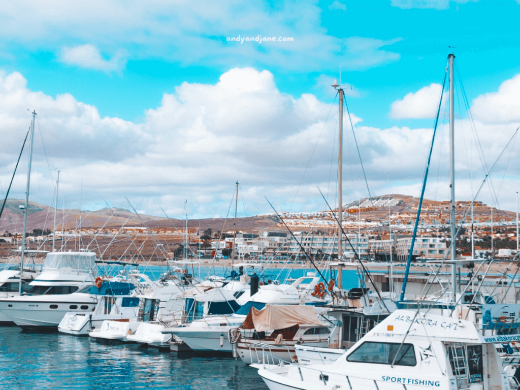 A bustling marina in Caleta de Fuste with various boats docked, against a backdrop of vibrant blue skies and clouds. Mountains and buildings are visible in the distance.