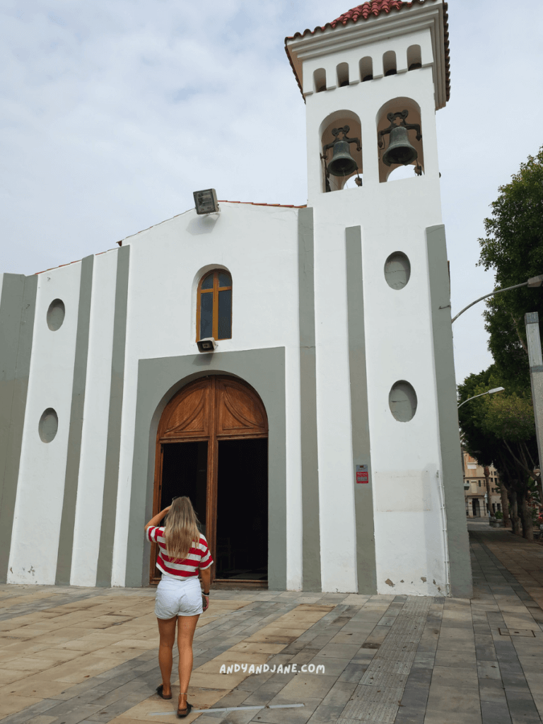 A woman in a striped shirt walks towards a white church with a red-tiled roof and bell tower on a cloudy day in Gran Tarajal, Fuerteventura