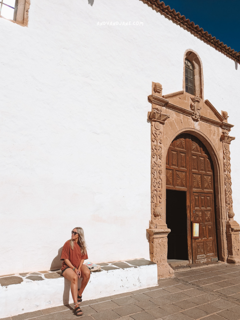 A person sits on a stone bench beside an ornate wooden door, in front of a whitewashed wall under a bright blue sky.