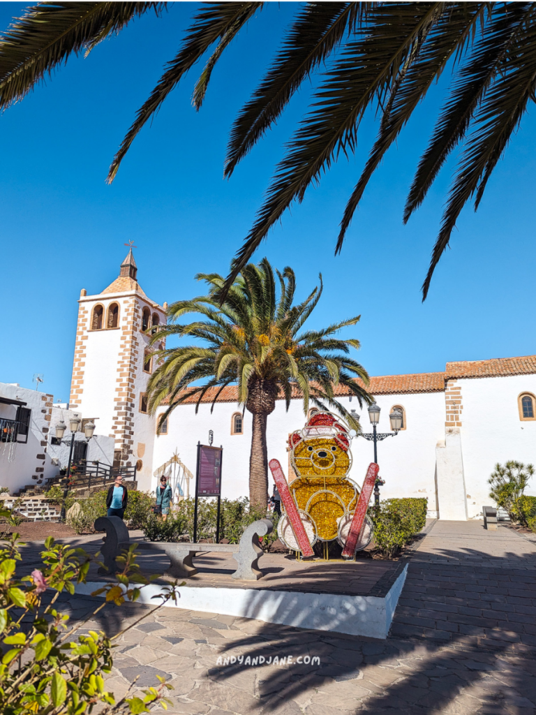 A colorful teddy bear decoration with ski poles stands amidst palm trees and a white church under a clear blue sky in Betancuria, Fuerteventura.