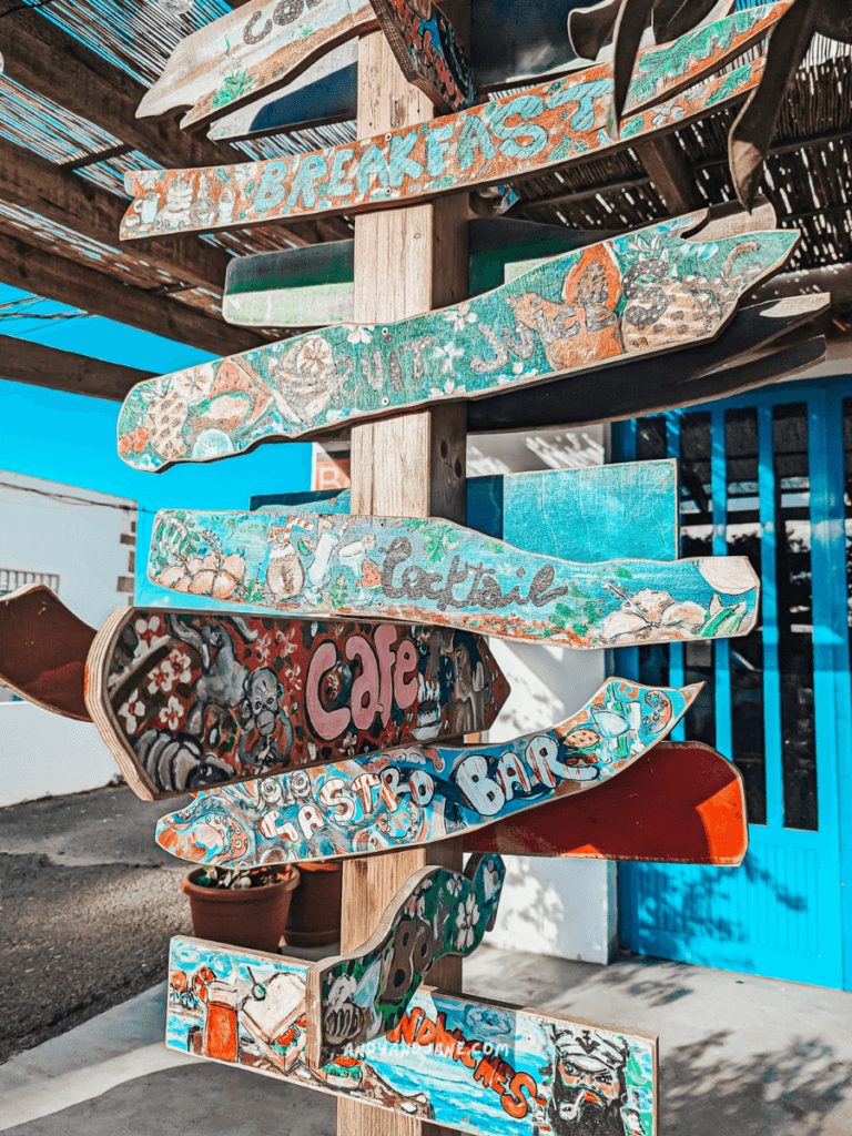 Colorful wooden signpost displaying various dining options like breakfast, cocktails, and a cafe, under a shaded awning in Lajares, Fuerteventura