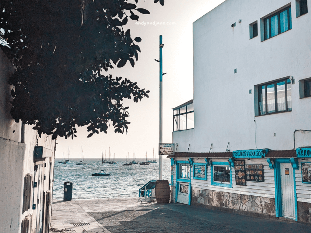 A charming seaside street in Corralejo with white buildings, a restaurant adorned in blue, and boats on the calm water in the background.