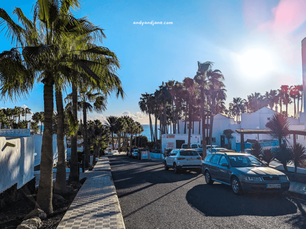 A sunlit street in Costa Calma, Fuerteventura, lined with palm trees and white buildings, featuring parked cars and a view of the ocean in the distance.