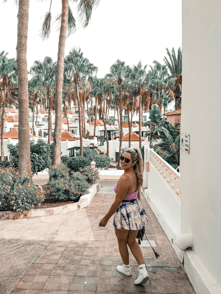 A woman in a colorful outfit strolls along a palm-lined pathway in Costa Calma, surrounded by lush greenery and white architecture.