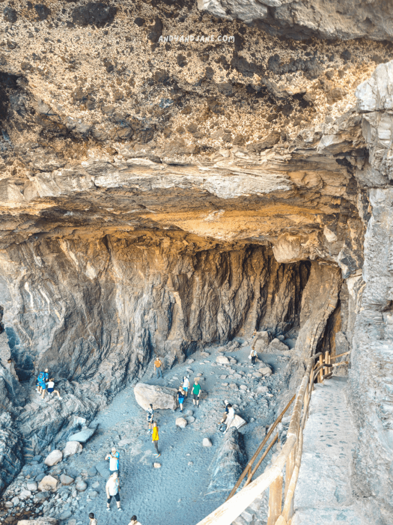 A panoramic view of Ajuy Caves with visitors exploring the sandy floor, surrounded by towering cliffs and large stones.