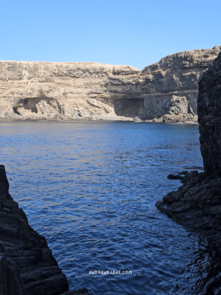 Serene coastal view at Ajuy Caves showing calm blue water between rocky cliffs under a clear blue sky, with sunlight illuminating the landscape.