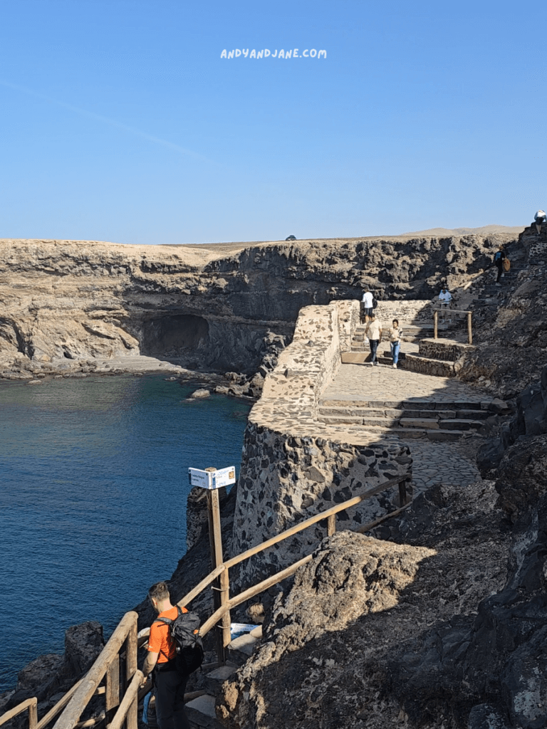 Walkers navigate a rocky coastal path with blue waters below and a clear sky above, surrounded by rugged cliffs.