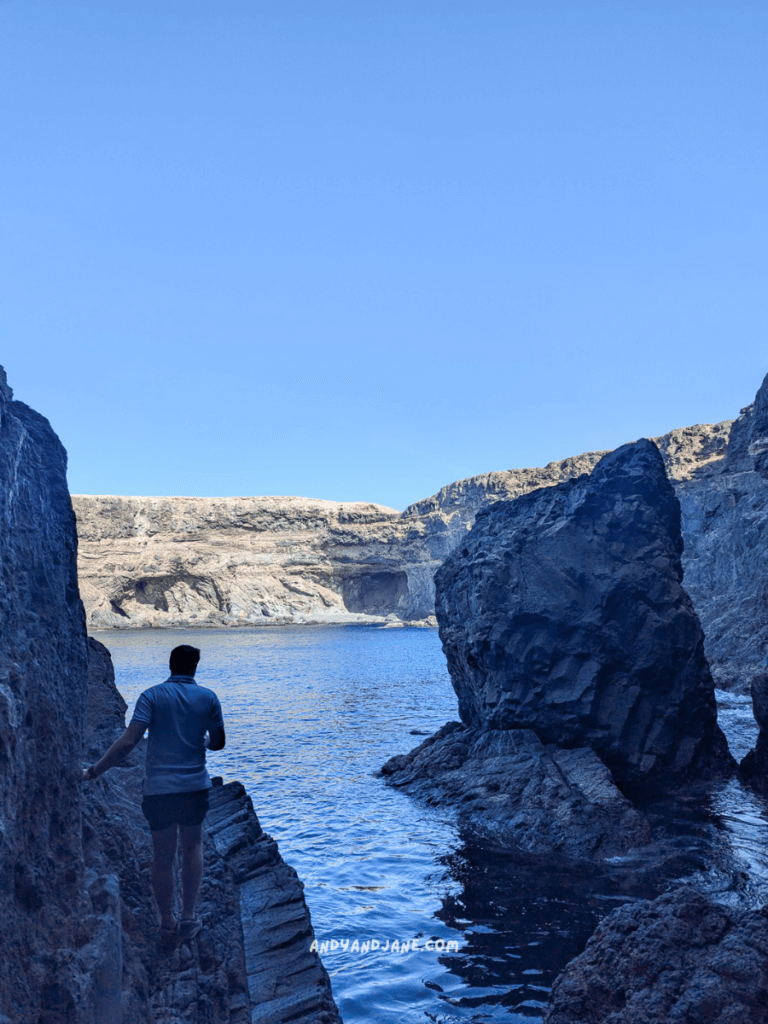 A person walks along rocky shores inside the Ajuy Caves beside calm blue waters, with cliffs and clear skies in the background.