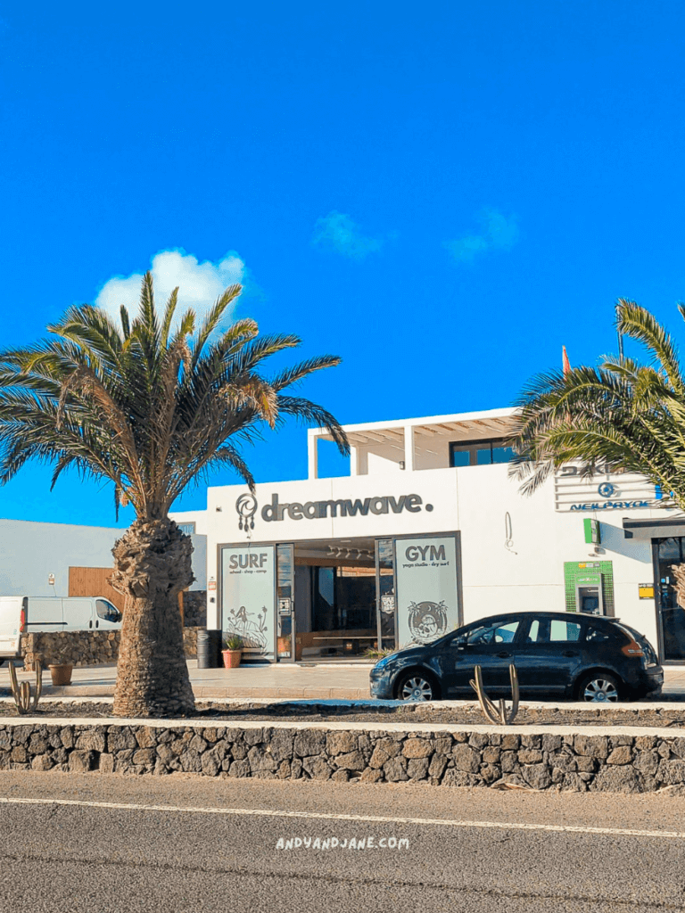 Exterior view of Dreamwave center featuring surf and gym facilities, flanked by palm trees under a bright blue sky.